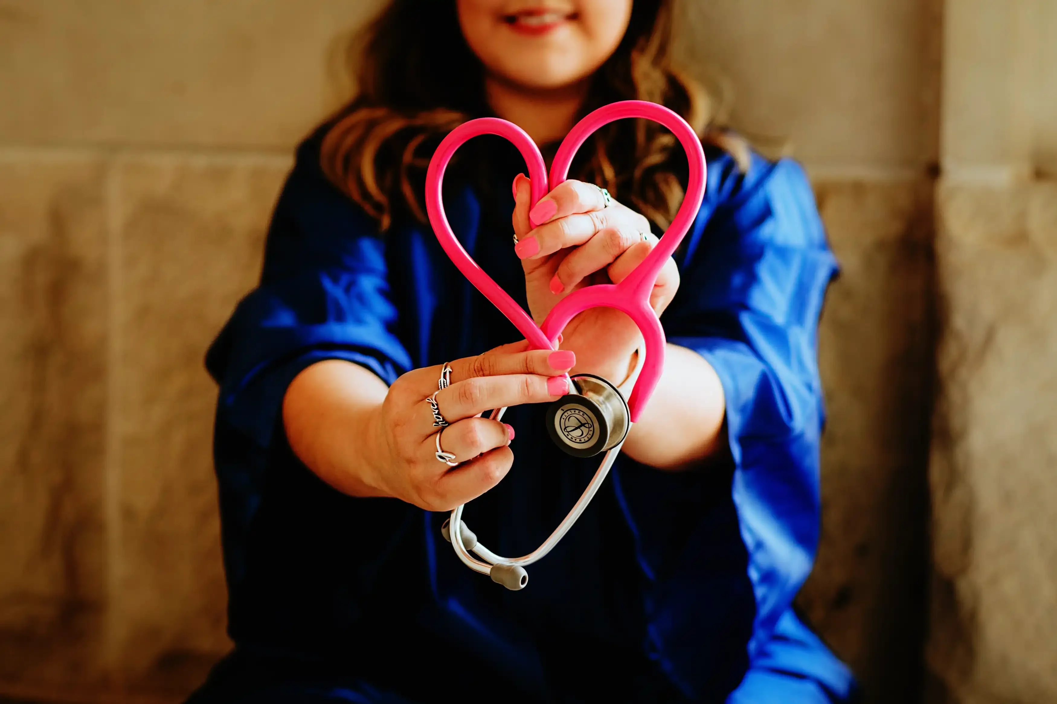A nurse in blue scrubs holding a pink stethoscope in the shape of a heart.